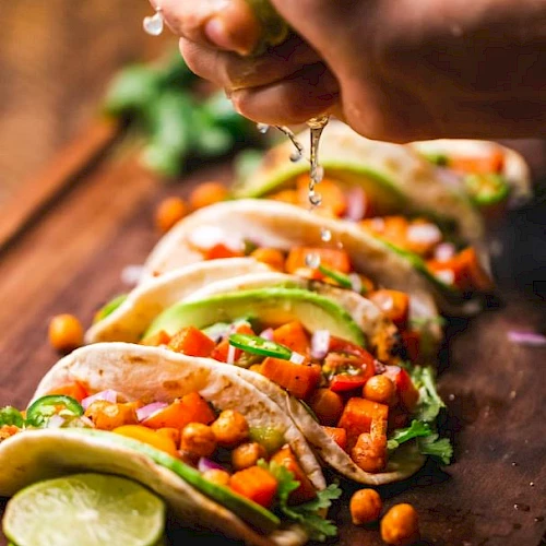 The image shows a hand squeezing lime juice onto a row of delicious tacos filled with chickpeas, avocado slices, cilantro, and diced vegetables.