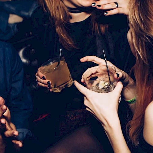 People are holding drinks and socializing in a lively atmosphere. One person is laughing while others engage in conversation with gestures.