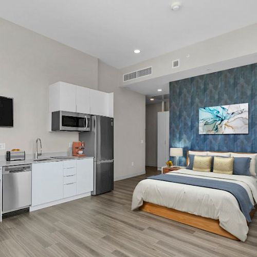 A modern studio apartment with a bed, kitchenette, desk, chair, TV, and abstract wall art on a blue accent wall.