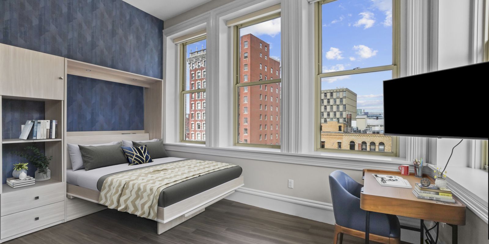 A modern bedroom with a large bed, desk, and chair facing a window with a city view. The room has wooden floors and a mounted TV on the wall.