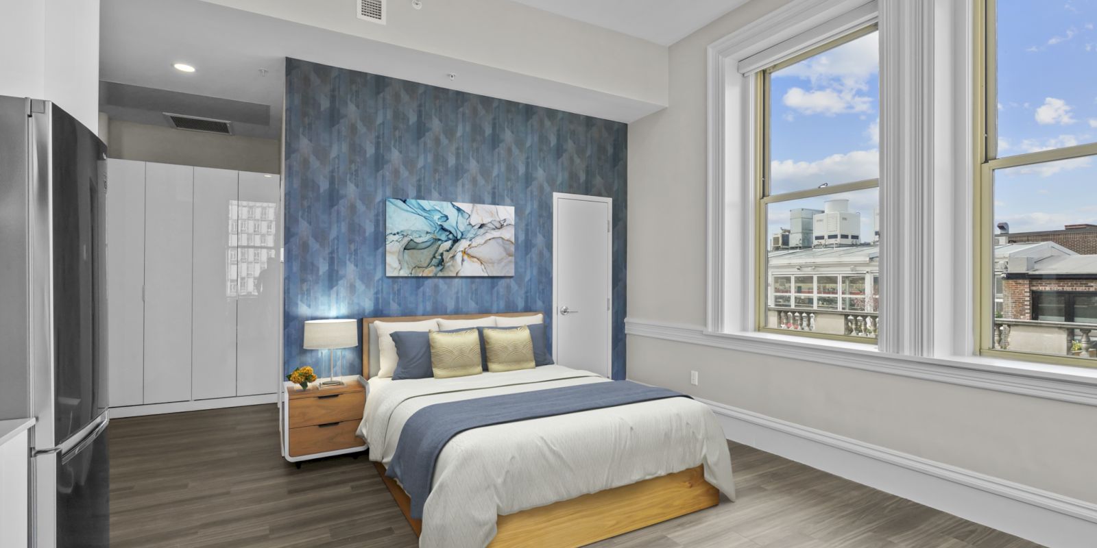 A modern bedroom with a bed, nightstand, and painting on the wall. Large windows offer city views and there is wood flooring throughout.