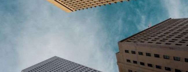 The image shows a dramatic upward angle view of tall skyscrapers against a blue sky with clouds, giving a sense of height and urban architecture.