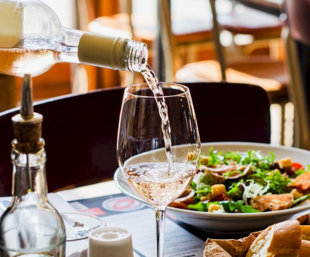 A bottle is pouring white wine into a glass on a restaurant table set with a salad, olive oil, bread, and condiments.