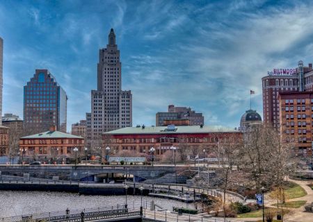 The image depicts an urban cityscape with several prominent buildings under a partly cloudy sky, featuring a river, park, and pedestrian bridges.