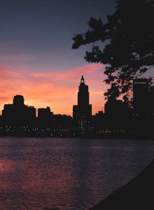 Silhouette of a city skyline at sunset with buildings, water, and a tree in the foreground. The sky has hues of pink and orange.