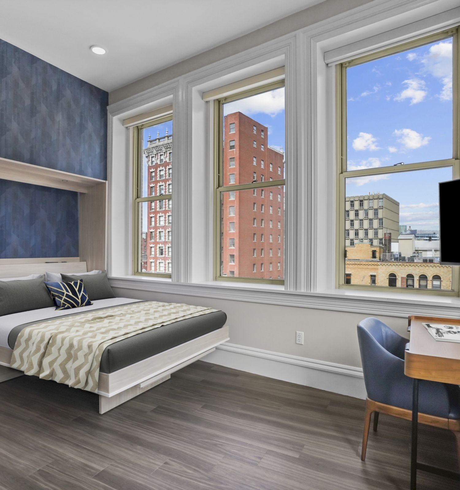 A modern bedroom with a large window view of buildings, a bed, a desk with a computer, and a chair. The room has wooden floors and blue accents.