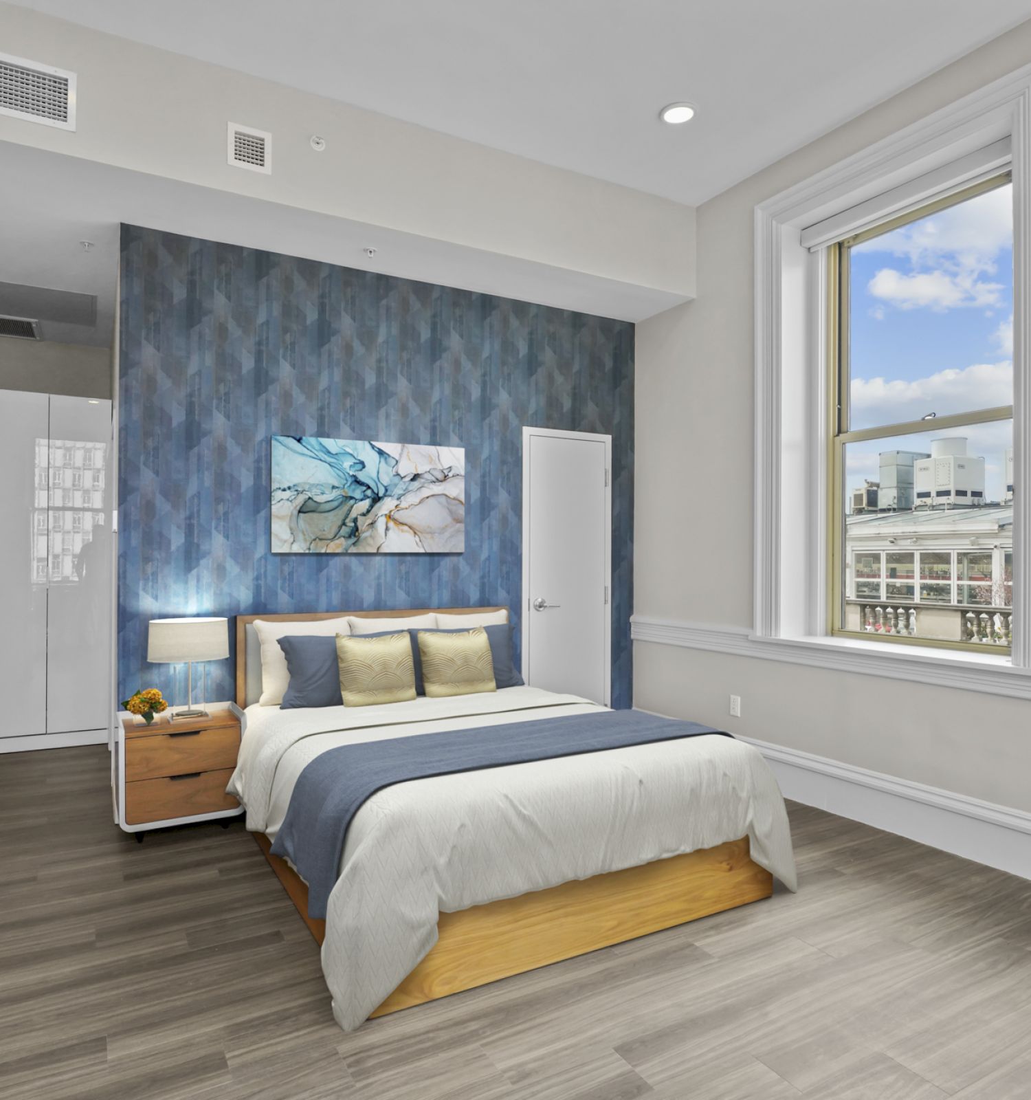 A modern bedroom with a large bed, blue accent wall, wardrobe, nightstand with lamp, wall art, and two large windows overlooking a cityscape ending the sentence.