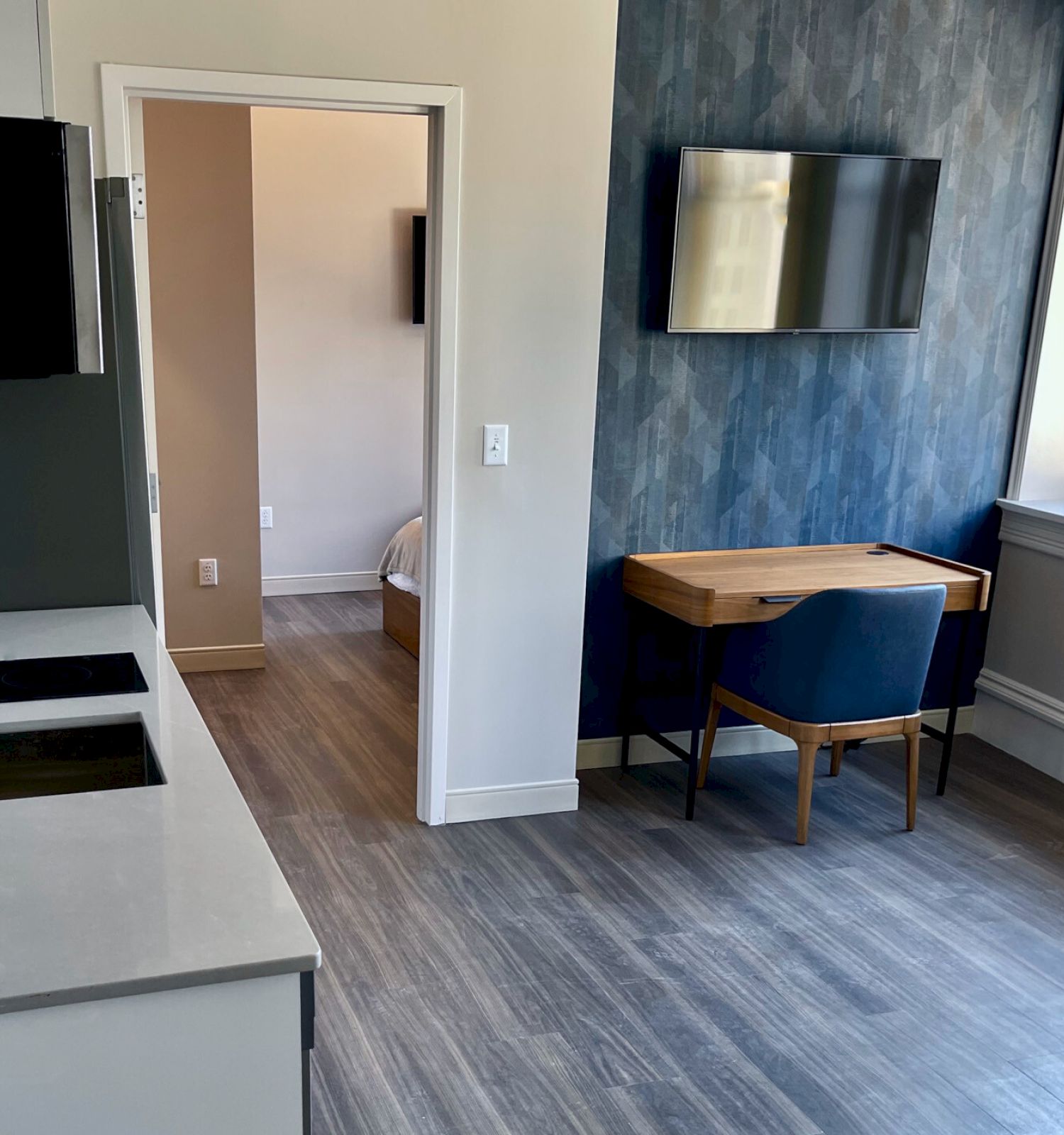 A modern room with a kitchenette, desk, wall-mounted TV, and large window; another room is visible through the door.