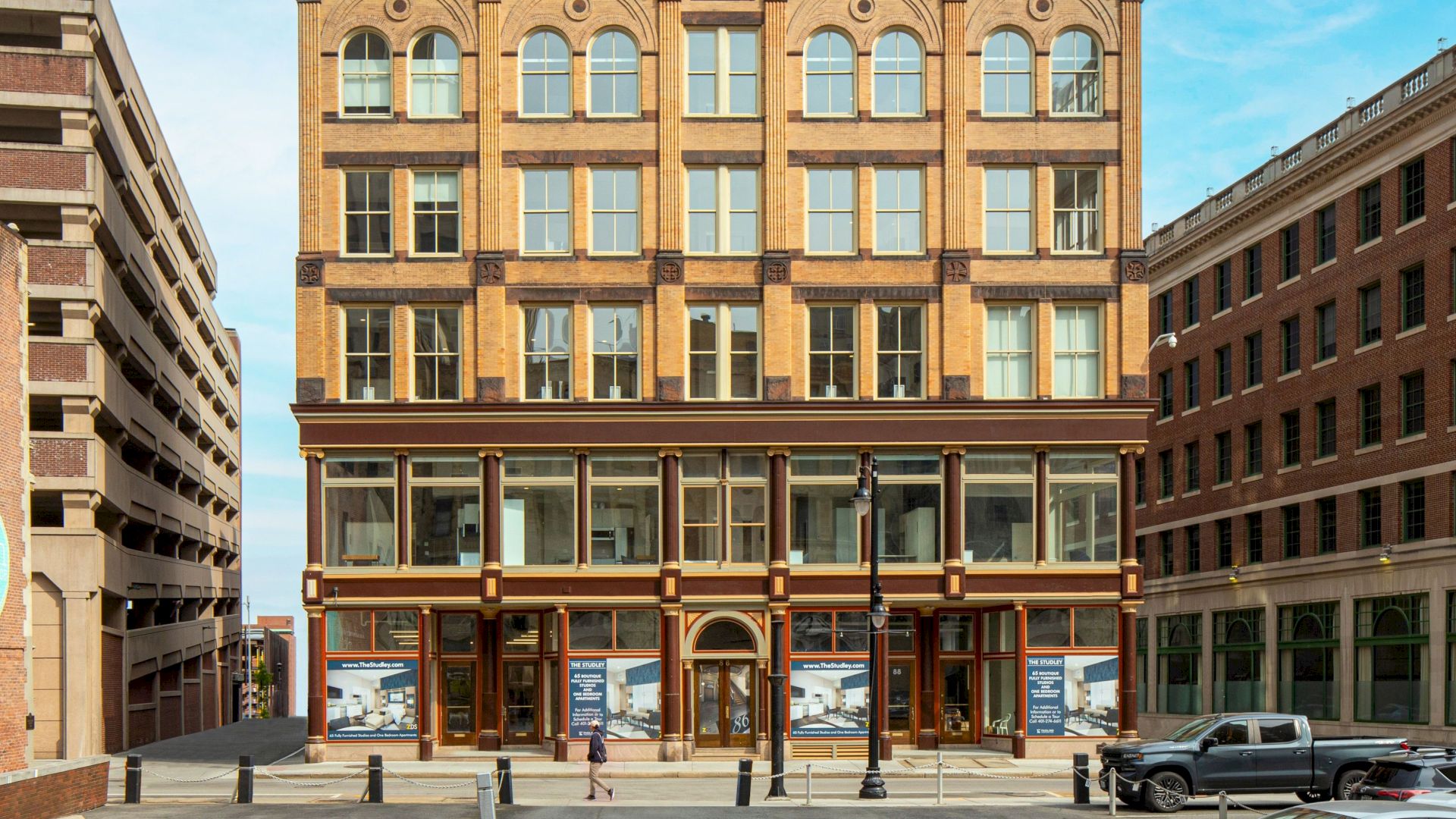 The image shows a multi-story brick building with large windows, flanked by a parking structure on the left and another building on the right.