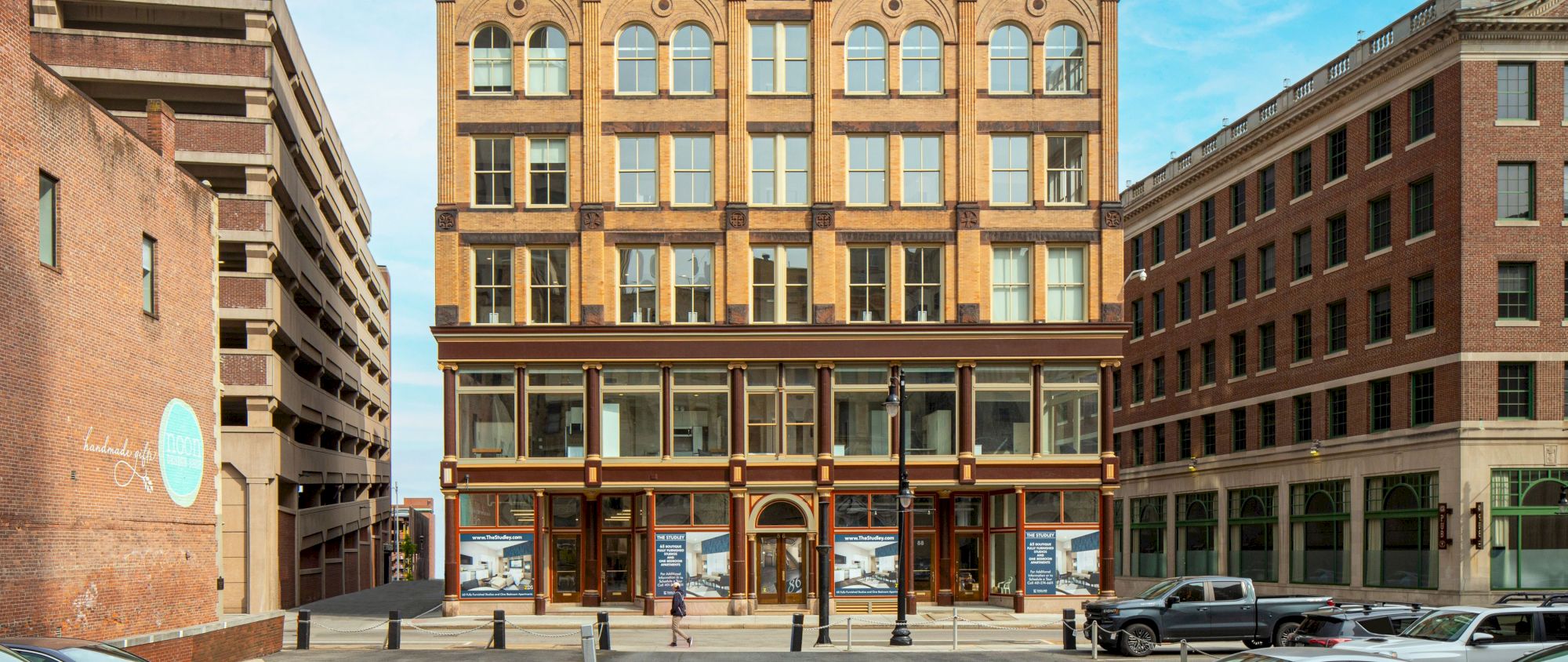 The image shows a multi-story brick building with large windows, flanked by a parking structure on the left and another building on the right.