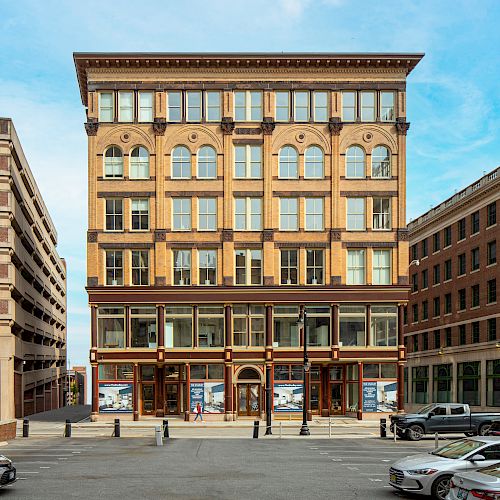 A historic multi-story building is centered between a parking lot and other buildings, featuring large windows and detailed architecture.