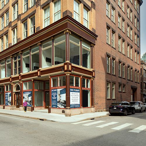 A street corner with a multi-story brick building featuring large windows, a crosswalk, parked cars, and a person walking on the sidewalk.