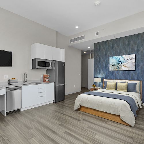 A modern studio apartment features a bed, kitchenette, desk, TV, and decor in a well-lit, spacious layout with wood flooring.