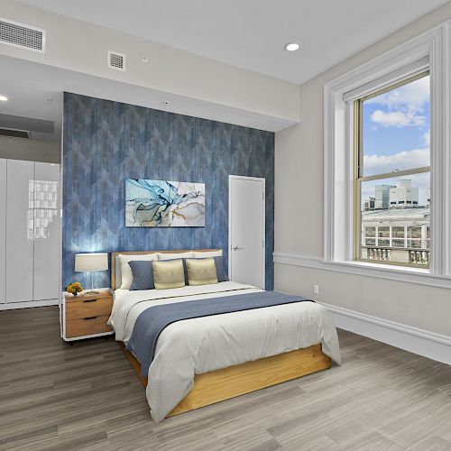 A modern bedroom with large windows, a double bed, nightstand, wall art, hardwood floor, and built-in storage in a well-lit space.