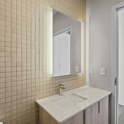 A modern bathroom with a large illuminated mirror above a sink, beige tiled wall, and open doorway to the right.