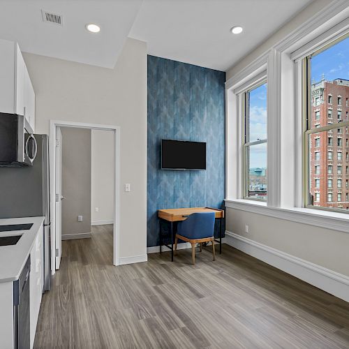 A modern kitchen and living space featuring large windows, a small dining area with two chairs, a wall-mounted TV, and wood flooring.