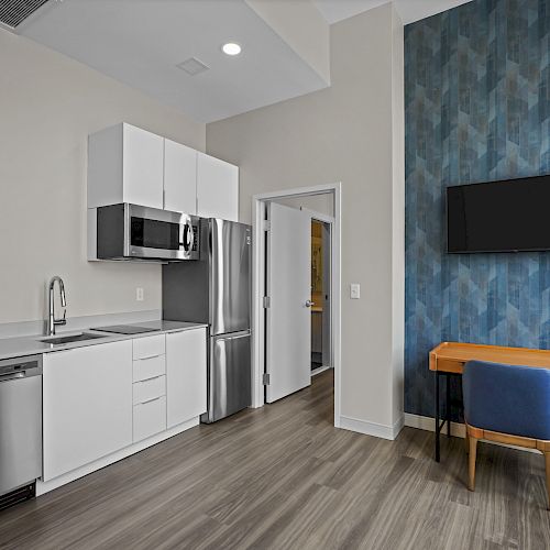 A modern kitchen with white cabinets, a microwave, stainless steel appliances, adjacent to a living area with a blue chair, desk, and TV.