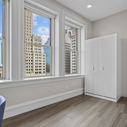 A room with three large windows, a white wardrobe, a blue chair, and a view of tall buildings outside.