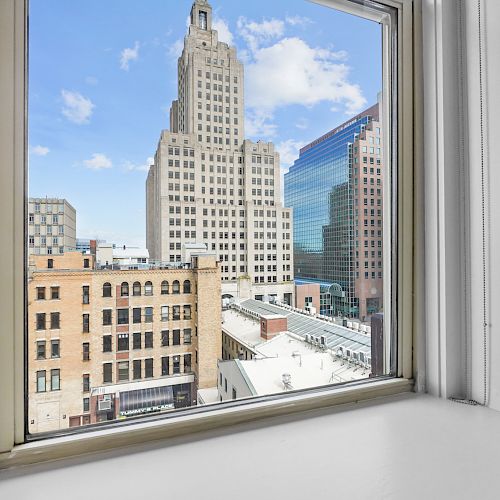 A cityscape view through a window, showcasing various buildings and a blue sky with some clouds.