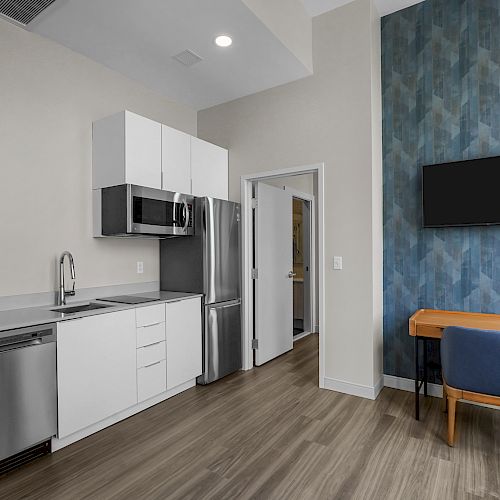 A modern kitchen features white cabinets, stainless steel appliances, and wood flooring; a nearby cozy workspace includes a desk, chair, and wall-mounted TV.