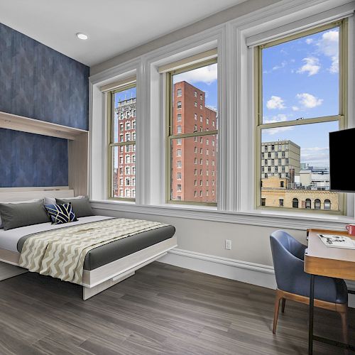 A modern bedroom with large windows, a bed with built-in storage, a desk and chair, and a flat-screen TV on the wall, with a city view outside.