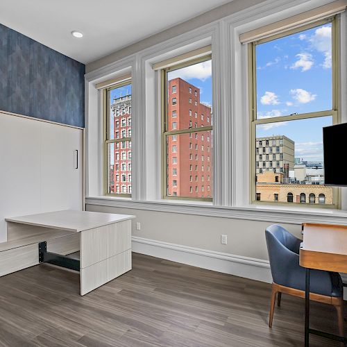 A modern office with a desk, chair, wall-mounted TV, large windows, blue accent wall, and cityscape view outside, featuring hardwood floors.