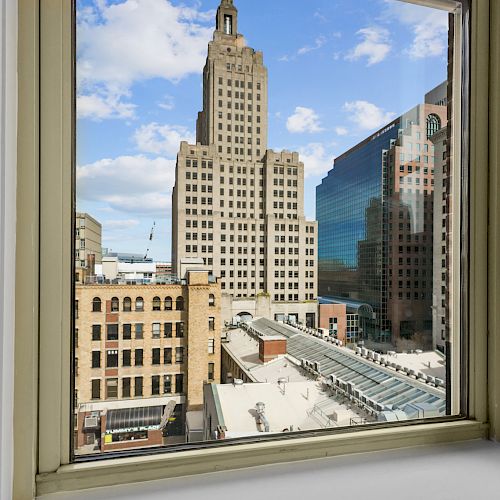 This image shows a cityscape framed by a window, featuring tall buildings under a partly cloudy sky, giving an urban view.