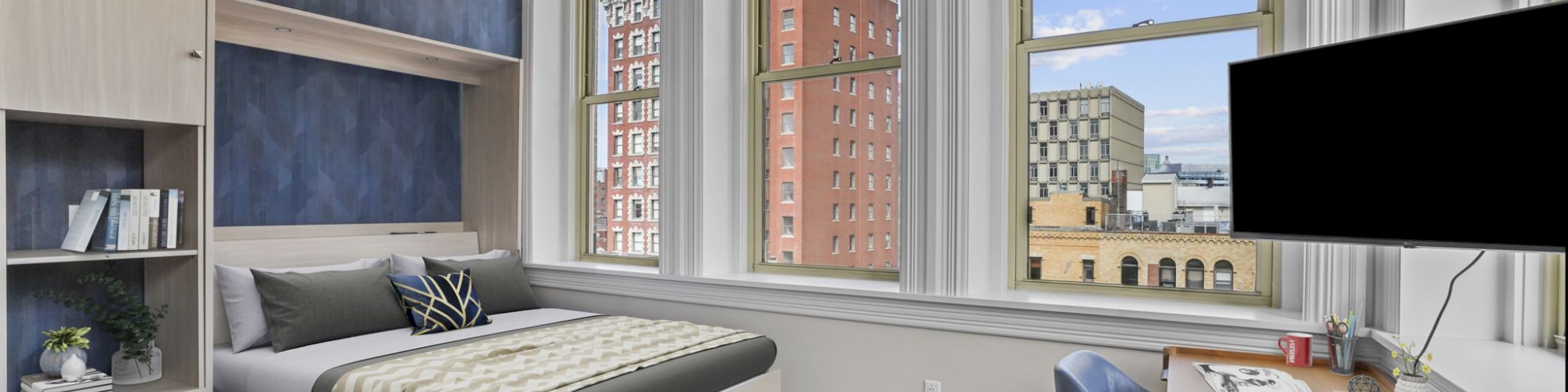 A modern bedroom with a bed, desk, and large windows showing a cityscape view. The room has wooden floors and contemporary decor.